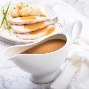 A gravy boat filled with turkey gravy and roast turkey breast and mashed cauliflower in the background.