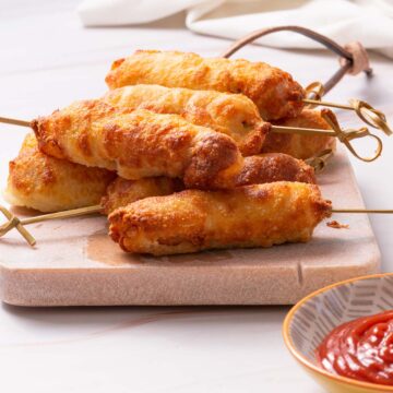 A stack of low carb corn dogs on a cutting board with a small bowl of ketchup.