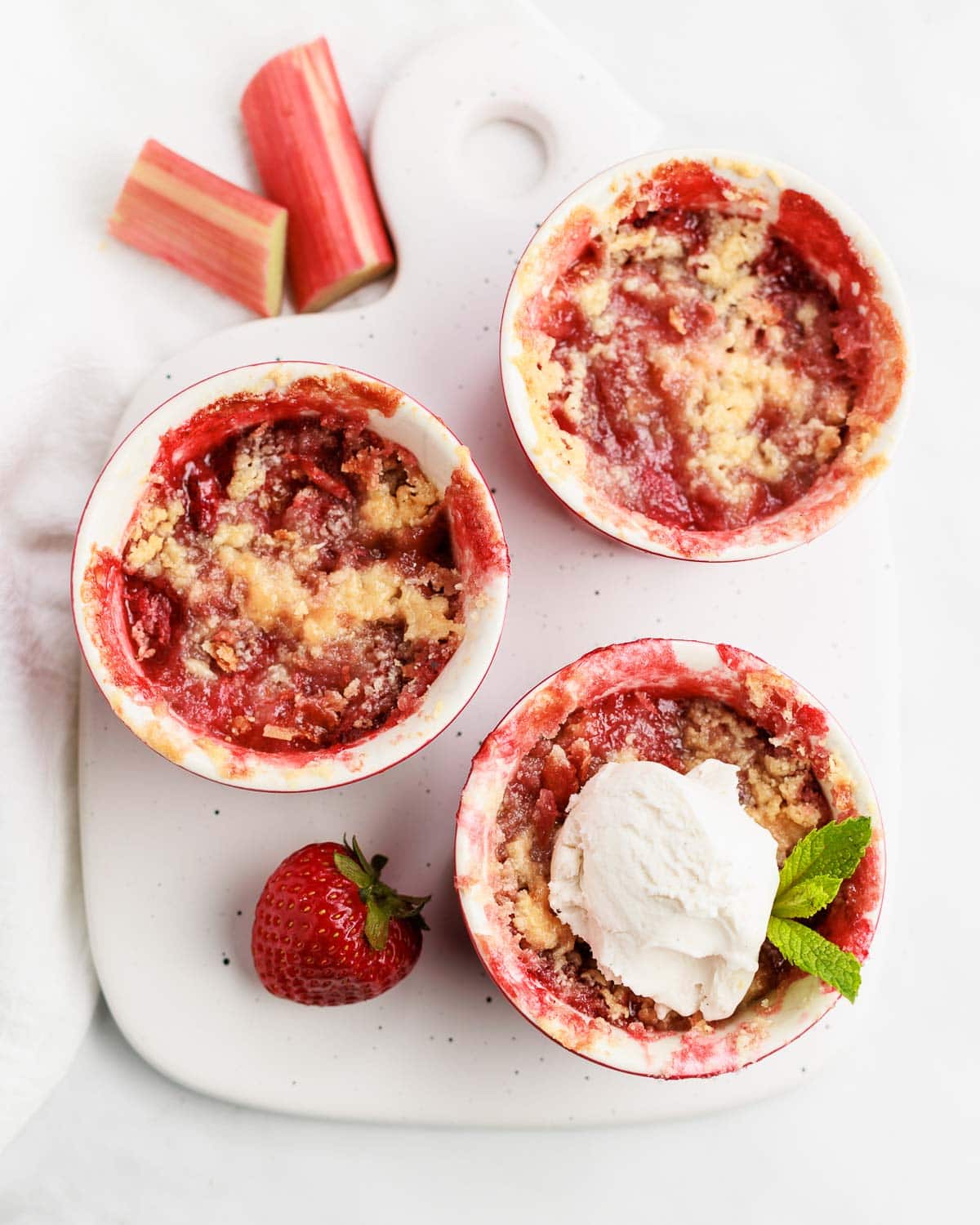 Three strawberry rhubarb crisps in a ramekin with  a scoop of ice cream on one .